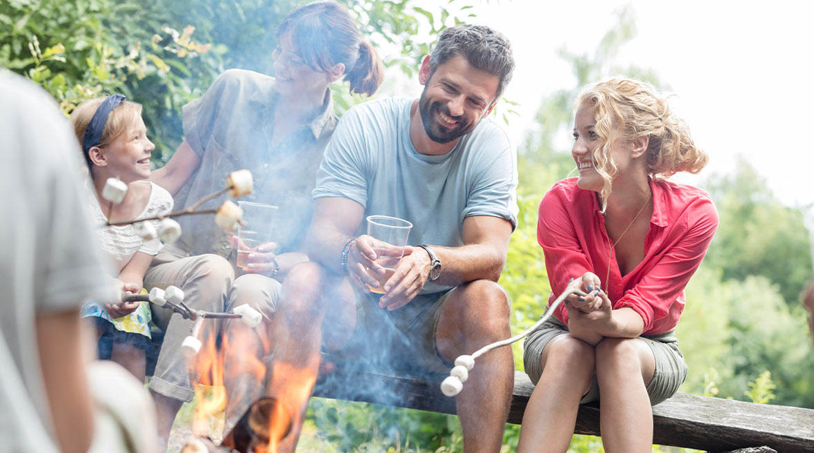 Family laughing and roasting marshmallows sitting on a log with blue sky in the background